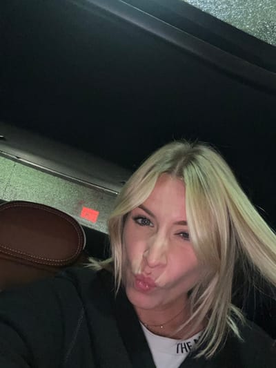 A selfie-style shot shows a blonde woman indoors, angled slightly. Her hair is windblown, and she is puckering her lips. She wears a white t-shirt with lettering on the upper right. A black hoodie covers the rest of her upper body. A brown car seat is visible below her. The ceiling is black. The image appears to be taken inside a car.