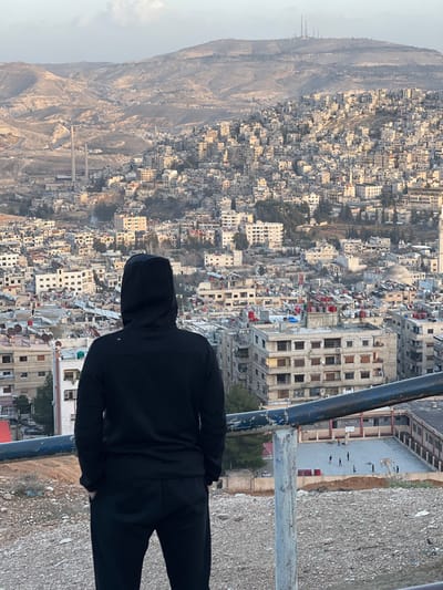 A single individual stands with their back to the camera, overlooking a densely populated urban area identified as Qudsaiya, Syria. The person wears a plain black hooded sweatshirt and black athletic pants, with hands seemingly in pockets. They are positioned behind a two-bar metal railing, painted blue and grey with visible rust, on a gravelly, elevated dirt patch. The midground reveals a vast expanse of multi-story residential and commercial buildings, predominantly light-colored concrete or stone, with numerous flat roofs often topped with water tanks or small structures. Scattered trees are visible among the buildings. To the lower right, a concrete sports court is discernible, featuring several small, dark figures, likely children, engaged in activity. The background depicts further urban expansion ascending the lower slopes of barren, rugged mountains. On the far left horizon, two tall, cylindrical industrial stacks are visible. On the right horizon, a prominent peak is crowned with multiple communication towers. The sky is pale and diffused, indicating either early morning or late afternoon conditions.
