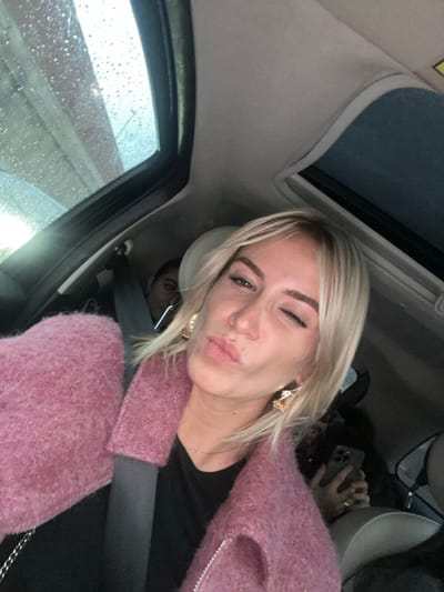 A casual selfie captures a young woman inside a car in Saint-Laurent-du-Var, France. She is the central subject, presenting a playful expression with a "duck face" and a wink of her left eye. She has blonde hair and light skin, and is wearing a soft, pink, fuzzy jacket over a black top. Large, ornate golden earrings adorn her ears.
The setting is the interior of a car, with a beige headliner and a visible sunroof above. A car seatbelt is fastened across her chest. Through the car window on the left, raindrops are visible, indicating wet weather. In the background, another individual is partially visible, holding a smartphone. Another person's head is faintly discernible behind the main woman, and a reflection of a face can be seen in her left eye. The image conveys a lighthearted and candid moment. No visible text is present.