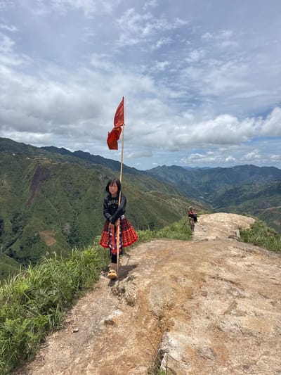 A young girl stands on a rocky overlook in Bắc Yên, Vietnam, holding a bamboo pole with a red flag. She is wearing a black sweatshirt and a traditional red and white patterned skirt. Behind her, the landscape unfolds into a vast panorama of lush green mountains, with terraced fields visible on the slopes. The sky is partly cloudy, with patches of blue peeking through. In the distance, another person, possibly an adult, is seated on the ground, with a child standing nearby. The terrain is rugged and uneven, with dry grass and rocks dotting the foreground. The overall scene depicts a remote, mountainous region with a hint of cultural tradition.