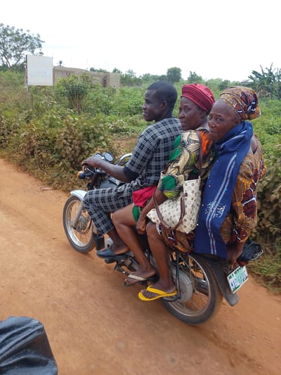 Three people, two women and a man, are riding on a motorcycle on a dirt road. The man is driving the motorcycle, wearing a black and white checkered shirt and pants. The two women are passengers behind him. The woman in the middle wears a red headscarf and a patterned dress. The woman in the back wears a colorful headscarf and a blue scarf around her neck, along with a patterned dress. Both women are looking towards the camera. They are all dressed in traditional African clothing.

The scene appears to be rural, with lush green vegetation on either side of the road. In the background, there is a partially constructed building and some trees. The sky is overcast, suggesting it might be a cloudy day. A license plate on the back of the motorcycle reads "OYO" and "MAP - 6420 G". A white handbag with a brown logo is visible on the woman in the middle's lap. The overall impression is of a typical mode of transportation in a Nigerian village.