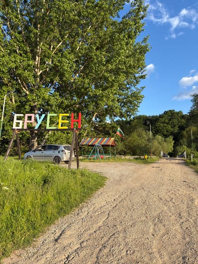 The image shows a dirt road leading into a forested area, presumably a park. On the left, there's a large tree and a sign that reads "БРУСЕН" (Brusen in Cyrillic alphabet) with each letter painted a different color. A parked car is near the sign. A colorful children's climbing structure is behind the sign. The flag of Bulgaria is atop the climbing structure. The sky is blue with some scattered clouds. Based on the sign "БРУСЕН" and context, this is in or near the village of Brusen, Bulgaria.