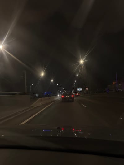 The image depicts a nighttime road scene captured from within a vehicle in Jilava, Romania. The dark sky is illuminated by numerous streetlights, which exhibit prominent starburst lens flare effects, lining both sides of a multi-lane asphalt road. White lane markings delineate the road surface. To the left, a concrete barrier separates the immediate roadway from an elevated structure or overpass. In the foreground, the top of a vehicle dashboard is visible, reflecting ambient light and the red taillights of traffic ahead. Ahead in the middle lane, a dark-colored sedan with illuminated red taillights is visible, followed by other vehicles further down the road, indicated by their red taillights. On the right side of the road, a blue rectangular road sign and a circular regulatory sign are discernible. A distinct blue light source is present in the distant left, and faint illuminated structures are visible in the distant right. No individuals are identifiable.