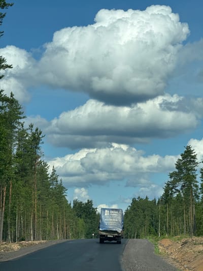 The image depicts a view from a vehicle traveling on an asphalt road through a forest area. The road is paved and has a visible shoulder. On either side of the road, there are dense forests composed primarily of tall pine and birch trees. The sky is blue with large, fluffy white clouds. In the distance, a blue truck with a white logo that reads "OKHHA PANORAMA" is visible, driving in the same direction as the viewer. The truck appears to be carrying goods covered by a blue tarp. The overall impression is a scenic drive through a natural landscape.