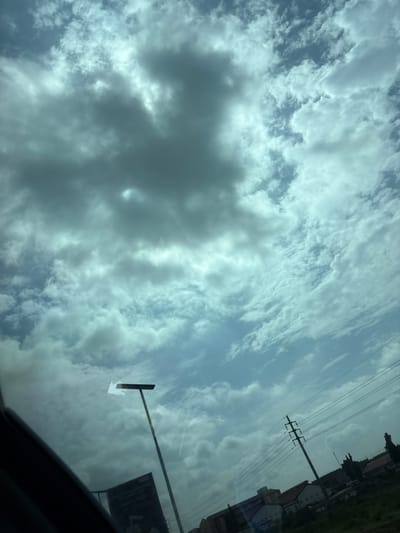 The image captures a wide-angle view of a cloudy sky over what appears to be a semi-urban or developing area in Gwaska, Nigeria. The sky is filled with a mix of dark, heavy clouds and lighter, wispy formations, suggesting potentially overcast or changing weather conditions. In the foreground, the bottom left corner shows a portion of what looks like the interior of a vehicle, possibly a car, indicating the photo was taken from inside.
Dominating the middle ground is a tall, dark lamppost with a horizontal fixture at the top, casting a faint reflection on the surface from which the photo is taken, likely a window. To the right of the lamppost, a large, dark billboard or sign is partially visible, its text obscured by the lighting and angle. Further to the right and in the background, power lines are strung between utility poles, indicating existing infrastructure. Below the power lines, a line of low-rise buildings and trees can be seen, some with red roofs. The overall impression is one of a typical day in a Nigerian city, with visible signs of development and everyday infrastructure.