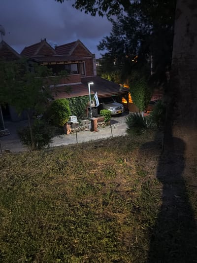 The image depicts a nighttime scene of a residential area in Yokneam Illit, Israel. A house with a red-tiled roof is visible on the left side of the frame. A parked silver car is near the front of the property. The area has dense vegetation and the lighting conditions are dark.