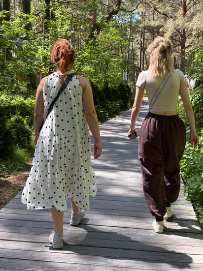 Two individuals walk away from the camera on a wooden boardwalk through a forest. The person on the left is wearing a white dress with black polka dots and a black and white patterned strap across their chest. They have reddish hair pulled back. The person on the right is wearing a light-colored crop top and dark brown sweatpants. They have blonde hair styled in two buns. The boardwalk is weathered and grey, surrounded by lush green trees and foliage, with sunlight filtering through the canopy, creating dappled shadows. The scene appears to be in a natural, outdoor environment.