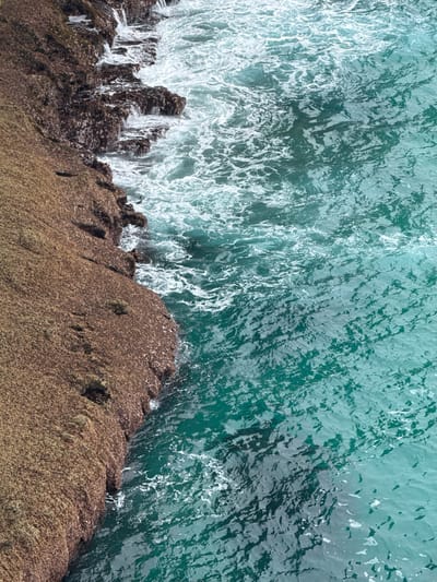 The image shows a high-angle view of a rocky coastline meeting the ocean. The rocks are reddish-brown and covered in a textured, granular material, suggesting they are weathered. The water is a vibrant turquoise, with white foam and waves crashing against the shore. The ocean surface is rippled, indicating moderate movement. There are no people or man-made objects visible. The scene is likely a natural coastal landscape, possibly in or around Llanes, Spain, known for its rugged coastlines.