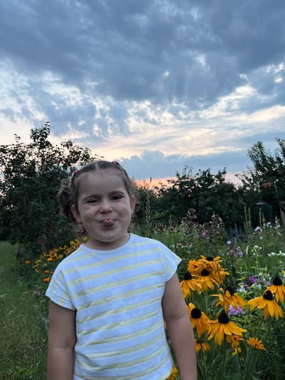 A young girl with her tongue sticking out stands in front of a garden. She has brown hair pulled into two pigtails. She is wearing a yellow and white striped t-shirt. The garden is filled with yellow and purple flowers and green foliage. In the background, there are trees and a cloudy sky with hints of sunset colors. The location is Elizówka, Poland.