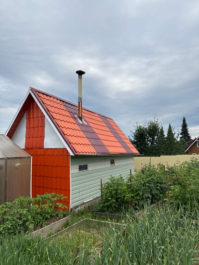 In the city of Lyady, Russia, a small, utilitarian building with a two-toned red and brown metal tiled roof is depicted. The building has a white gable end and red siding on the lower half of its front and side. A metal chimney extends vertically from the roof. To the left of the building is a small, translucent greenhouse. The foreground of the image shows a garden with rows of green plants, including what appears to be green onions or chives and potato plants. In the background, a light yellow fence separates the garden from a denser area of trees, with another small, wooden house visible in the distance on the right. The sky is overcast with gray clouds.