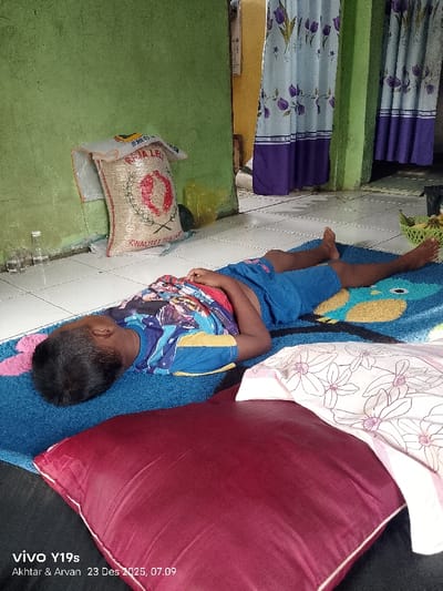 A young boy is sleeping on a blue mat with a cartoon design. He wears a blue t-shirt and shorts. A large red pillow sits near his head. Behind him, a sack of rice leans against a green wall. Curtains with a tulip pattern hang in the background. A basket of bananas rests on the tiled floor near his feet. Bottles are visible in the distance near the sack of rice. A watermark is at the bottom-left of the image.