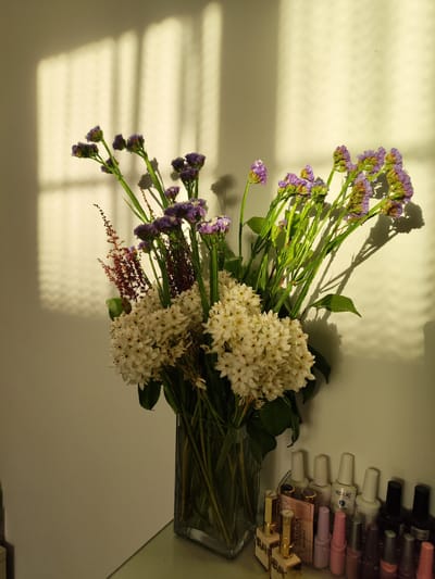 A still life photograph features a bouquet of flowers in a clear glass vase, placed on a surface with a row of nail polish bottles arranged in front of a light-colored wall. The flowers are a mix of white and purple, with some reddish-brown sprigs. Sunlight casts long shadows of window blinds onto the wall behind the flowers, creating a patterned effect. The nail polish bottles are of various colors and brands, including Rosalind. The scene is set indoors, likely in a domestic environment, as suggested by the presence of nail polish. The location is identified as Pico da Pedra, Portugal. There are no people or animals visible in the image. The overall lighting suggests late afternoon sun.