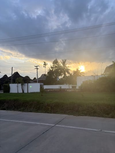 The image depicts a scene in Ban Ton Kwen, Thailand, during a sunset. The sky is filled with dramatic clouds, with the sun peeking through, casting a warm golden light. Several houses with distinctive architectural styles, featuring dark, steeply pitched roofs, are visible in the background. These structures appear to be modern homes or villas, some with white walls. In the foreground, there is a well-maintained grassy area bordered by a white wall. A paved road with white markings runs across the bottom of the frame, suggesting a street or thoroughfare. Electrical poles and wires are also present, indicating the presence of infrastructure in the area. The overall atmosphere is serene and natural, characteristic of a tropical sunset.