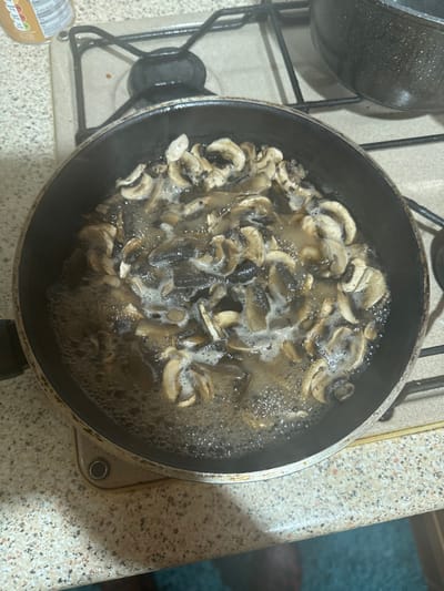 A black non-stick frying pan, positioned on a gas hob with visible metal pan supports, contains a quantity of sliced mushrooms immersed in a vigorously bubbling, light-brown liquid. The mushrooms are varied in shade from light to dark brown. To the left of the hob, a light-colored, speckled countertop is present. In the upper background, a dark cooking pot and a partially visible food container with text are discernible. A faint blue-green surface is visible below the countertop. The scene depicts a culinary process occurring within a kitchen environment in Fowlis Easter, United Kingdom.