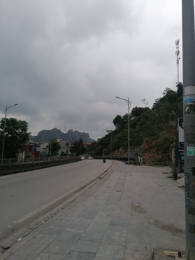 The image depicts a street scene in Ha Tu Ward, Vietnam, on an overcast day. A gray concrete road curves gently to the left, bordered by a concrete barrier and a gray tiled sidewalk. A motorcycle is traveling on the road. In the distance are buildings and a mountain range. A telephone tower is visible on top of a hill to the right of the road. The right side of the sidewalk is overgrown with vegetation. A utility pole is on the right of the frame. The sky is filled with heavy gray clouds.