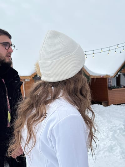 The scene is set outdoors in a snowy environment in Yeghipatrush, Armenia. In the immediate foreground, the back of a person is visible, featuring long, wavy light brown hair extending from beneath a white ribbed knit beanie. This individual is wearing a white jacket with a dark stylized symbol on the upper left back. To the left, partially visible in profile, is another individual, appearing male, wearing dark-rimmed glasses and possessing a beard. This person is dressed in a dark coat over a patterned garment. Both individuals are standing on ground entirely covered by white snow. In the background, two wooden structures resembling market stalls or small huts are present, with their roofs also covered in snow. A string of illuminated warm-toned light bulbs is suspended horizontally across the upper background, above the structures. The sky is overcast and grey.