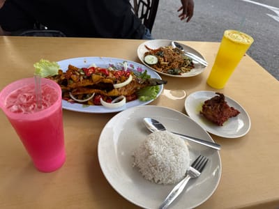 The scene depicts a casual meal laid out on a light-colored table at an outdoor eatery in Malacca City, Malaysia. In the upper left, a person wearing a black t-shirt is partially visible, seated at the table. A dark-skinned hand is also visible in the upper right background.

On the table, several dishes and beverages are arranged. A large oval light-blue plate, patterned with subtle florals, prominently features a whole grilled fish, likely sambal-grilled or sweet and sour style, topped with sliced red chilies and white onions, resting on a bed of lettuce. To the right, another oval white plate holds a stir-fried dish, possibly noodles or vegetables, garnished with a cucumber slice; a spoon and fork are placed on this plate. Below these, a plain off-white circular plate contains a mound of white rice, accompanied by a fork and a spoon. Adjacent to the rice, a smaller oval white plate holds a single piece of deep-fried chicken.

Two beverages are present: a tall plastic cup on the left contains a pink, iced drink with a straw, and a tall plastic cup on the right holds a yellow, iced drink with a straw. A water condensation ring is visible on the tabletop near the fish plate. The background reveals an asphalt surface, indicating an outdoor or roadside dining environment consistent with street food vendors or casual restaurants common in Malacca City.