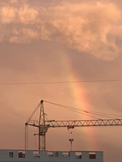 A construction crane is silhouetted against a soft, peachy sky, partially obscured by clouds, with a faint rainbow arching through the frame. The crane's yellow metal structure is clearly visible, with cables and a hook hanging down. Below the crane, the top of a brick building under construction is visible, with open window frames. The scene appears to be from Kaluga, Russia. There are no people visible in the image.