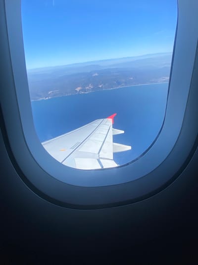 The image is a view from an airplane window looking out over a body of water and a coastline. In the foreground, the wing of the airplane is visible, with its characteristic white surface and red wingtip. The wing appears to be a commercial airliner, as suggested by its size and design. The water is a deep blue, likely the ocean, and it meets a rugged coastline with hills and mountains in the distance. The sky is clear and blue, indicating good weather. The image is taken from a high altitude, providing a panoramic view of the landscape. There are no people or discernible actions visible in the scene. The location is unknown, but the landscape suggests a Mediterranean coast, potentially in Spain, given the rocky terrain and clear blue waters.