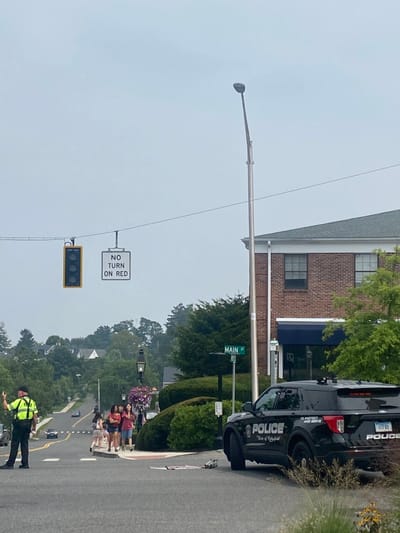 The image depicts a daytime street scene on Main Street in Ridgefield, United States, under an overcast sky. In the foreground-left, a male police officer, wearing a lime green reflective vest over a dark uniform and a dark cap, is standing with his right arm raised, signaling. A black Ford Explorer police SUV, marked "POLICE Town of Ridgefield" and bearing Connecticut license plate "117 RT," is parked on the right side of the roadway, facing away from the camera. Directly in front of the police vehicle on the pavement, a flattened cardboard box-like object and a grey cylindrical object are visible.
In the mid-ground, a group of four young female pedestrians are walking on the sidewalk from the background towards the viewer's left. Overhead, a traffic light displaying a yellow signal hangs alongside a "NO TURN ON RED" sign. A green street sign reading "MAIN ST" is visible on a pole on the right side of the road. A two-story brick building with white trim and a blue awning is located on the right. The environment includes mature trees, manicured bushes, and decorative street lamps, one featuring hanging baskets with purple flowers. Other residential structures are visible in the background, situated on a rising slope.