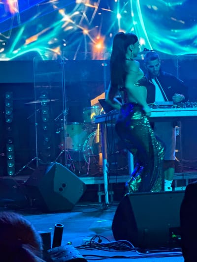 This image depicts a nighttime outdoor stage performance. A woman with long dark hair pulled into a ponytail, wearing a sequined outfit, is positioned in the foreground, her back to the camera, facing a DJ and a music setup. She appears to be in motion, possibly dancing. Behind her, a man with short dark hair, wearing a dark jacket over a white shirt, is seen operating a DJ console. The stage is illuminated by vibrant blue and green lights, with a large, abstract light display in the background. Several speakers are visible on the stage and in the foreground, along with drum equipment and cables. The setting suggests a concert or festival atmosphere. There is no information in the image that places the scene in Akhalubani, Georgia.