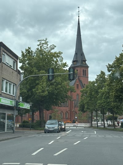 This is a street-level, full-frame, medium-distance, eye-level, overcast day, color photograph taken in Gronau, Germany. The main focal point is a tall, red brick church with a dark, pointed steeple topped by a weather vane. The church has arched windows and a clock on its facade. In the foreground, there is a paved road with white lane markings. Several cars are present on the road, including a light blue Mercedes-Benz A-Class and a dark colored car behind it. On the left side of the image, there is a building with a brick facade and windows. A traffic light is visible hanging over the intersection. Trees with green foliage line the street. In the background, there appears to be some construction activity with an excavator and fencing. A few pedestrians are also visible in the distance. The sky is cloudy and gray.