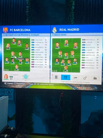 This image depicts a television screen displaying a soccer video game's team selection interface, set indoors in the city of Dustlik, Uzbekistan.
The screen is vertically divided. The left half showcases the FC Barcelona lineup in a 4-2-4 formation, featuring player portraits and ratings. Key players visible include L. Suárez (92), L. Messi (93), Iniesta (79), and O. Dembélé (79). A white cursor is positioned over "Sergio!" (87). A partial list of substitutes, including J. Cillessen (81) and P. Coutinho (86), is displayed to the right of the formation. Russian text above reads "Стартовый состав/Изменить позицию" (Starting lineup/Change position).
The right half presents the Real Madrid lineup in a 4-3-3 formation, also with player portraits and ratings. Prominent players shown are C. Ronaldo (94), K. Benzema (87), G. Bale (90), and T. Kroos (88). A partial list of substitutes, including Kiko Casilla (81) and Isco (87), is adjacent to the formation. Russian text above reads "Главное меню" (Main menu).
Below the team displays, game control prompts in Russian, such as "Справка" (Help), "Подтвердить" (Confirm), and "Назад" (Back), are visible. The lower section of the screen reflects the surrounding room, revealing dark green decorative elements and a partially visible "NO ALCOHOL" sign with a crossed-out beer bottle icon on the bottom left.