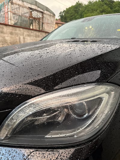 The image is a close-up of a black car, with water droplets covering the exterior. The car's headlight is prominent in the foreground. Reflected in the car's surface are glimpses of a building under construction, covered in scaffolding and netting. Trees are also visible in the background. The image does not provide any definitive information about its location being Zhaltusha, Bulgaria.