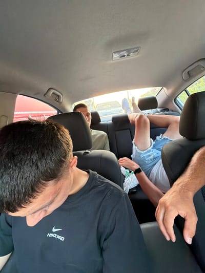 The media file shows the interior of a car from the perspective of a passenger. Three individuals are visible. In the foreground, a person is wearing a black t-shirt with a white Nike Pro logo. Their head is turned to the side, obscuring most of their face. Behind them, another person is seated, wearing a white t-shirt and ripped denim shorts, with their legs extended. A third person is visible further back in the car, appearing to be asleep or resting their head on the seat. The car's interior, including the headrests and ceiling, is gray. Sunlight streams in through the windows, suggesting it is daytime. The overall mood appears relaxed, possibly indicating a road trip or travel. The location is Giurgiu, Romania.