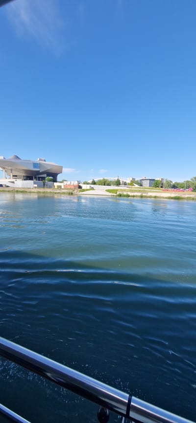 This image captures a panoramic view of the Rhône River in La Mulatière, France, under a clear blue sky. In the foreground, a polished metal railing, likely from a boat or observation deck, extends across the bottom of the frame, partially submerged in the dark blue-green water. The river exhibits distinct ripples and waves, reflecting the sunlight. The opposite riverbank displays a combination of modern architecture and urban development. On the far left, the distinct, futuristic design of the Musée des Confluences is visible, characterized by its metallic, undulating rooflines and glass facades. Moving right along the bank, there are additional buildings, areas of green foliage, and a white arched bridge spanning part of the landscape. A prominent pink sign or sculpture spelling "ONLY LYON" with a red lion emblem is visible on the right side of the riverbank. The sky, occupying the upper two-thirds of the image, is a bright, unclouded blue, with only faint, wispy cirrus clouds barely discernible in the upper left. The scene is brightly illuminated by direct sunlight.