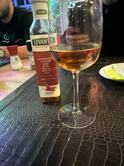 The image shows a wine glass containing a small amount of amber-colored liquid, lying on its side on a dark, textured surface resembling crocodile skin. Next to the glass is a bottle of Levante brand olive oil infused with chili peppers, its label clearly visible. Part of a person's face and wrist with a watch are visible in the lower left corner. The background appears to be the interior of a restaurant. There is no information about the location in the city of La Louvière, Belgium.
