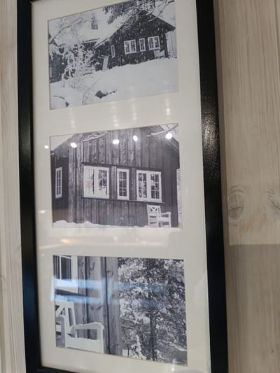 This media file contains a framed triptych of black and white photographs. Each photograph depicts a rustic, wooden building, likely a cabin or a house, in a snowy environment. The overall mood is serene and quiet, suggesting a winter scene.

The top photograph shows a larger structure with multiple windows, partially covered by snow. A snow-laden tree or bush is prominent in the foreground. The building appears to have a dark, possibly shingled roof.

The middle photograph offers a closer view of a section of a wooden building, featuring several windows. One window on the left is a single pane, while the central and right windows are multi-paned. A white wooden chair or bench is placed outside one of the windows. Icicles are visible hanging from the roofline.

The bottom photograph is a split image. The left side shows a white wooden bench or chair similar to the one in the middle photo, set against a dark, vertical wooden wall. The right side of the bottom image focuses on a snow-covered tree with intricate branches, characteristic of a winter landscape.

The photographs are presented in a mat with a white border, framed by a black frame. The background against which the framed images are displayed appears to be a light-colored, textured wooden wall, possibly paneling.

Based on the provided location context, these images likely depict scenes from Hovden, Norway, a popular Norwegian ski resort known for its winter sports and natural beauty. The weather depicted is clearly cold and snowy, characteristic of a Norwegian winter. No people are visible in the photographs, and there is no visible text.