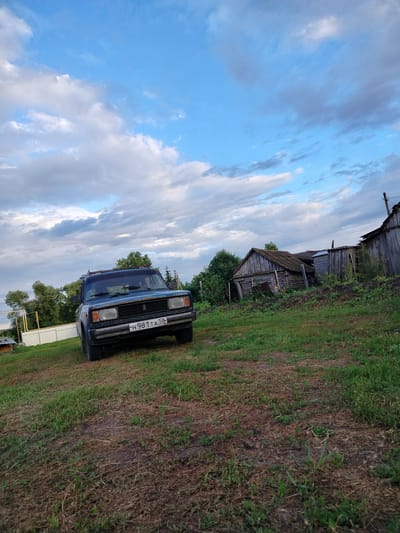 The media file depicts an outdoor scene in what appears to be a rural setting. In the foreground, a blue, older model car is parked on a grassy area. The car has a roof rack and a license plate that reads "H 981 TA 58 RUSSIA". Behind the car, there are several dilapidated wooden structures, resembling sheds or small houses, with weathered roofs and walls. Green trees and foliage are visible throughout the scene, suggesting a natural environment. The sky above is partly cloudy with a mix of blue sky and gray clouds, indicating it might be late afternoon or early evening, possibly before or after a rain shower. The overall mood of the image is somewhat rustic and possibly melancholic due to the state of the buildings and the overcast sky. The location is identified as Вазерки, Russia. There are no people visible in the media file.