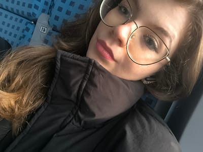 A close-up, angled portrait captures a female subject from the upper torso upward, situated within what appears to be a passenger vehicle. The subject has light brown hair and is wearing gold-framed, cat-eye style eyeglasses over light-colored eyes. Her lips are painted with a dark pink lipstick. She wears a black puffer jacket with a high collar. A dark earbud or hearing aid is visible in her right ear. The background features a blue upholstered seat with a repeating square pattern, and a partially visible white tag marked with "A" and a stylized upward arrow, indicative of a seat identifier. This scene is located in the city of Kopanitsa, Bulgaria.