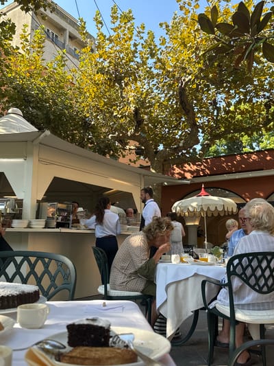 This image depicts an outdoor dining area in Sestri Levante, Italy, under a clear blue sky, beneath the canopy of large trees with green and yellowing foliage.
In the left-to-center, a white, multi-peaked outdoor serving station is visible, containing stainless steel kitchen equipment and stacks of white plates. Two staff members are positioned behind the counter: a female with dark hair in a ponytail, wearing a white shirt and dark trousers, appearing to arrange items; and a male with dark hair tied back, wearing a white shirt and dark apron, standing near the female. Another individual wearing a light-colored cap is partially visible behind the counter.
Multiple patrons are seated at white-clothed tables with dark green metal chairs, some with white cushions. In the mid-ground, an elderly woman with blonde, curly hair, wearing a patterned long-sleeved top, is actively eating from a cup with a spoon. She is seated opposite an elderly man with gray hair and glasses, wearing a light blue collared shirt, and an elderly woman in a white patterned top. A young blonde child is partially visible between the two elderly patrons, seated at the same table. This table holds white cups, plates, and glasses containing orange liquid.
In the foreground, a separate table displays a round dark chocolate cake dusted with powdered sugar. Another plate on this table contains a slice of similar dark chocolate cake and a piece of light-brown flat pastry.
On the right side of the scene, a decorative white umbrella with a red finial and scalloped trim provides shade over an additional seating area, with a terracotta-colored wall casting shadows behind it.