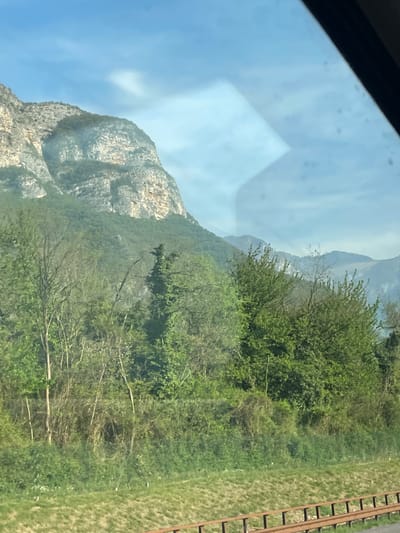 A daytime shot, taken from inside a vehicle, looks out onto a landscape in Rivalta, Italy. To the left is a large, gray, rocky mountain under a blue sky with light clouds. Green trees and foliage cover the lower slopes of the mountain and extend into the foreground. To the right is a strip of green grass alongside rusty railroad tracks. The scene is viewed through a slightly dirty car window, as evidenced by the smudges and glare. The photo appears to have been taken while the vehicle was in motion.