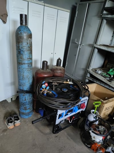 The image depicts a storage area within a workshop or industrial facility, possibly located in Shimanovsk, Russia. In the foreground, a portable generator is prominent, connected to a set of coiled hoses and welding equipment. To the left of the generator are two large gas cylinders, one tall and blue, the other shorter and red. Behind these, white lockers and a tall grey metal cabinet are visible, suggesting a structured storage system. A pair of worn athletic shoes rests on the concrete floor near the base of the blue cylinder. The overall environment appears utilitarian, with evidence of ongoing work or stored equipment.