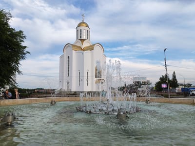 A white, domed Orthodox church with a golden cupola stands prominently in the background, adorned with a cross at its apex. In the foreground, a multi-tiered fountain sprays water, creating a dynamic display. Two people are visible near the fountain's edge on the left, and another person can be seen near the church entrance.

The scene appears to be a public square or park, with a tree to the left and a street with some buildings and vehicles visible behind the church. The sky is partly cloudy, suggesting it might be late afternoon or early evening. There is a lamppost with a sign on it to the right of the church, and a digital billboard in the distance. Some people are gathered on a walkway behind the fountain. The overall mood is peaceful and somewhat tranquil, with the water feature adding a lively element to the static architecture. The presence of the church and the style of architecture are consistent with locations in Kurgan, Russia. No specific text is clearly legible from the image.