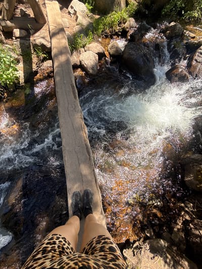 An overhead, eye-level shot shows a person's legs and feet standing on a narrow wooden log bridge spanning a rocky stream in Allenspark, Colorado. The person is wearing black sneakers with black socks and a leopard-print skirt. The stream is fast-flowing with white water cascading over dark rocks. Sunlight glints off the water's surface. The surrounding environment includes large rocks and green vegetation. The wooden bridge appears weathered and rustic. The location is a natural outdoor setting, likely a hiking trail or park area within or near the city of Allenspark.