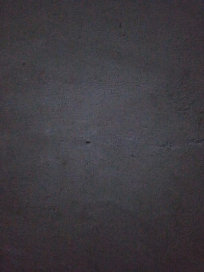 A dark, mottled gray surface, possibly concrete or asphalt, dominates the image. Its texture appears coarse and uneven, with subtle variations in shade suggesting roughness. Dim lighting obscures fine details, creating a somber and muted tone. A few small, dark specks are scattered across the surface, adding to the visual interest of the texture.