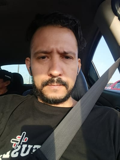 A man with dark, wavy hair, a beard, and a mustache is depicted in a close-up, self-portrait style photograph taken inside a car. He appears to be in his 30s or 40s, wearing a black long-sleeved t-shirt adorned with a prominent white and red graphic; partially visible letters include "US" or "GUS" with a stylized cross-like emblem above. Blue wireless earbuds are in both his ears. A grey seatbelt crosses his chest diagonally, indicating he is seated in a moving or parked vehicle. His expression is neutral, almost contemplative, with a slight furrow in his brow.
Bright sunlight illuminates the scene from the left, casting distinct shadows on his face and shirt, suggesting a clear day, likely morning or late afternoon. Through the car's rear window, a light blue sky and the faint outlines of buildings or structures are visible. In the background, another individual is partially seen in the back seat, wearing a dark cap with reversed white text that reads "HAQROL." A small portion of a red car is also visible through the right side window. The car interior suggests an everyday urban or suburban setting, consistent with Rímac, Peru.