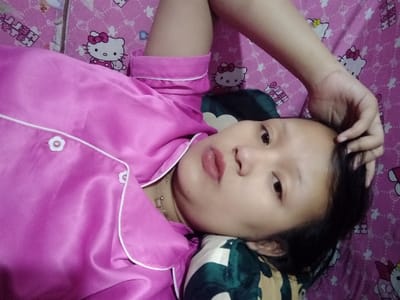 A person lies on a pink Hello Kitty bedspread, wearing a pink pajama top with white trim. They are resting their head on a pillow, with their hand near their forehead. They are looking slightly off-center towards the camera. A gold necklace is visible at their neckline. The scene is lit with soft, indirect light.