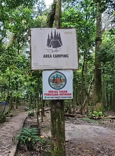The image shows a signpost in a forest setting, likely a campsite. The signpost is made of wood and has two signs attached to it. The top sign is white with a drawing of a tent and trees, and the text "AREA CAMPING" below it. There is also some smaller text at the bottom of this sign. The second sign, mounted below the first, is a circular logo with a tent and mountains, and the text "SHARLTON CAMP" above it. Below the logo, in red text, are the words "KERJAS TAKAN PENATAJAN OUTDOOR". At the very bottom of this second sign, there are social media handles "@HATLLLINE TSNO" and "@AREAOUTDOOR". The background consists of tall trees with lush green foliage, indicating a natural environment. A path made of stones or concrete is visible to the left of the signpost, winding through the forest. The ground is covered with fallen leaves and dirt. The lighting suggests it is daytime, with diffused sunlight filtering through the trees. The scene appears to be in a relatively natural and possibly remote area, consistent with a camping ground. The location is identified as Dolat Rayat, Indonesia.
