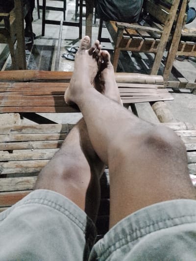 The media file shows a close-up view of a person's legs and feet, resting on a wooden bench. The subject is wearing light-colored shorts. The background consists of a dimly lit outdoor setting with wooden furniture, possibly part of a seating area. In the distance, a pair of flip-flops are visible on the ground. The lighting suggests it could be evening or nighttime. The overall mood is relaxed and casual. There is no visible text in the image. The setting appears to be outdoors in a tropical or subtropical location, possibly a park or a casual eatery, given the simple wooden furniture and the presence of flip-flops. The location context provided is Sugihwaras, Indonesia.