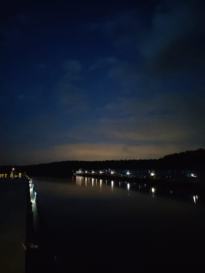 The image depicts a waterfront scene at night in Ermolino, Russia. A dark body of water reflects the lights from a row of buildings on the opposite shore. Silhouetted trees line the horizon behind the buildings. A pier extends into the water on the left side of the frame. The sky is a gradient of dark blue with cloud cover. No people are visible.