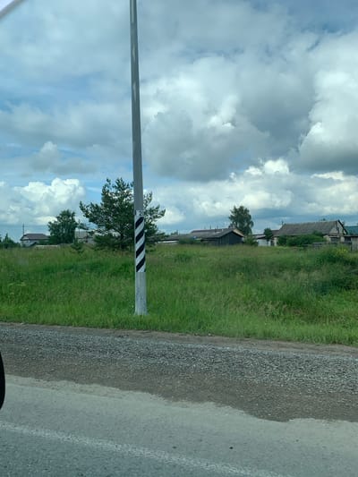 The image shows a roadside view in the village of Nevolino, Russia. In the foreground, a gravel and asphalt road is visible. A gray utility pole with black and white diagonal stripes and a marker indicating "2" stands prominently in the middle ground. Behind the pole, a field of tall green grass stretches across the scene, dotted with wildflowers. Several rustic wooden houses with gray roofs are scattered among the trees and vegetation in the background. The sky is partly cloudy with large, dramatic cumulus clouds against a blue backdrop. A single pine tree and other deciduous trees are interspersed throughout the landscape.
