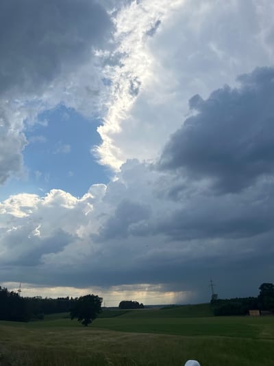 The image captures a dramatic sky over a rural landscape in Tuntenhausen, Germany. Dark, ominous storm clouds dominate the upper portion of the frame, with shafts of sunlight breaking through in the distance, creating an ethereal glow. Below the turbulent sky, a green, rolling field stretches towards a dark treeline. A solitary tree stands in the middle ground, silhouetted against the lighter sky. In the far distance, a power line pylon and a small building are visible. The overall impression is one of an approaching or receding storm, casting a moody and atmospheric light on the pastoral scenery. There are no people or identifiable animals visible in the frame. The image appears to be a landscape photograph taken from a slightly elevated vantage point.