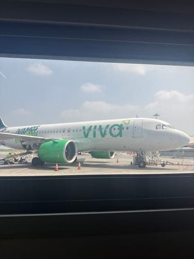 The image shows a Viva Aerobus airplane viewed through a window. The plane is white with green accents, parked on an airport tarmac. In the background, are other airport structures and a cloudy sky. The photo was likely taken in Apodaca, Mexico.