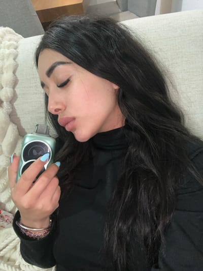 A woman lies on a beige couch with dark hair, wearing a black top. She holds a light green electronic device in her right hand. Her fingernails are painted a light blue. She has a bracelet on her wrist. Her eyes are closed, and she appears to be resting. The background is a soft, fuzzy blanket.