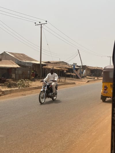 The image depicts an urban street scene in a location within Nigeria. A male individual, wearing a light-colored long-sleeved tunic, light trousers, and a small cap, is riding a black and chrome motorcycle with a prominent round headlight. He is positioned in the left lane of an asphalt road, moving from left to right across the frame. On the far right, the rear and right side of a yellow three-wheeled auto-rickshaw is partially visible. The roadside on the left features a row of single-story buildings, many with corrugated metal roofs and walls, appearing as commercial or residential structures. Two smaller figures are visible near a building on the left. The sky is hazy and light, with multiple utility poles and a dense network of overhead power lines visible against it. The verges alongside the road are unpaved, consisting of bare earth.